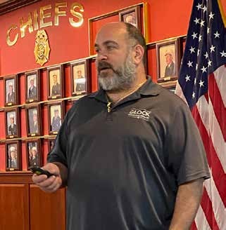 Jeff Hirschman standing in front of a wall of framed photos and an American flag, answering student questions during a concealed carry license class.